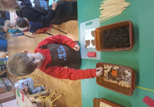 dzieci ubrane w brązowe fartuszki biorą udział w czekoladowych warsztatach, samodzielnie ozdabiają czekoladę w kształcie serca