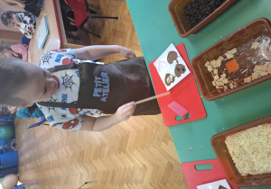 dzieci ubrane w brązowe fartuszki biorą udział w czekoladowych warsztatach, samodzielnie ozdabiają czekoladę w kształcie serca