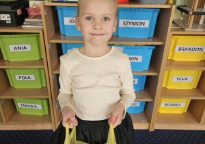 Dziewczynka w czapeczce urodzinowej stoi, trzymając w rękach reklamówkę.