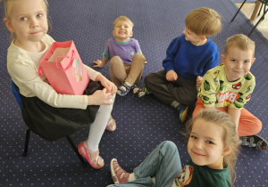 Dziewczynka siedzi na krześle, a dzieci siedzą na dywanie.