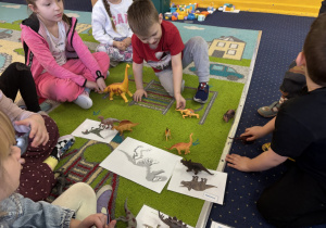 Dzieci siedzą w kole na dywanie i oglądają figurki dinozaurów.