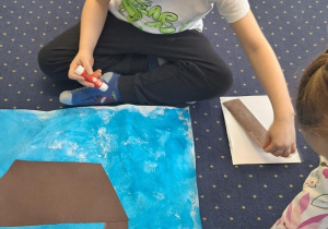 Dzieci siedzą na dywanie i wykonują wspólną pracę plastyczną.