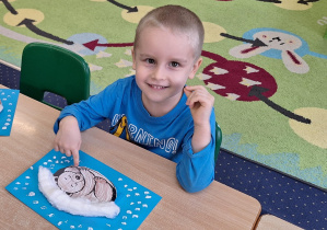 Chłopiec siedzi przy stole i pokazuje swoją pracę plastyczną.