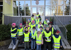 grupa dzieci ubrana w żółte, odblaskowe kamizelki pozuje do zdjęcia przed wejściem do szkoły