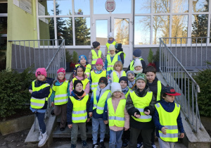 grupa dzieci ubrana w żółte, odblaskowe kamizelki pozuje do zdjęcia przed wejściem do szkoły