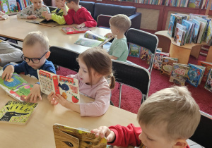 Dzieci siedzą przy stolikach i oglądają książki.