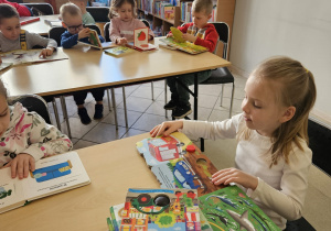Dzieci siedzą przy stolikach i oglądają książki.
