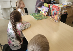 Dzieci siedzą przy stolikach i słuchają pani.