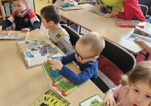 Dzieci siedzą przy stolikach i oglądają książki.