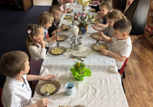 Dzieci siedzą przy stołach i zjadają wielkanocny obiad.