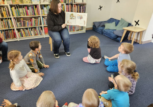 Dzieci siedzą na dywanie i słuchają książki czytanej przez panią.