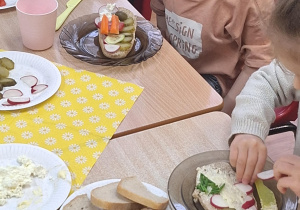 Dzieci siedzą przy stole i zjadają samodzielnie przygotowane śniadanie.