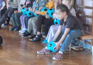 Dzieci siedzą na ławeczce z piłkami.