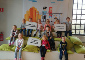 Dzieci z grupy 1 siedzą na podeście i troje z nich trzyma w dłoniach hasła związane z Łodzią.