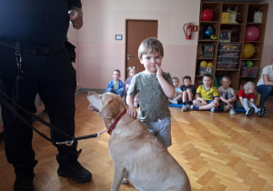 chłopiec głaszcze psa w obecności strażnika miejskiego, na sali gimnastycznej w tle widać siedzące dzieci