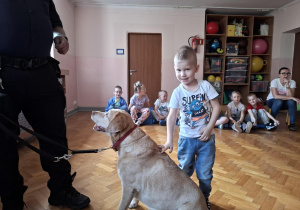 chłopiec głaszcze psa w obecności strażnika miejskiego, na sali gimnastycznej w tle widać siedzące dzieci