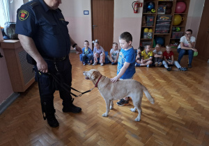 chłopiec głaszcze psa w obecności strażnika miejskiego, na sali gimnastycznej w tle widać siedzące dzieci