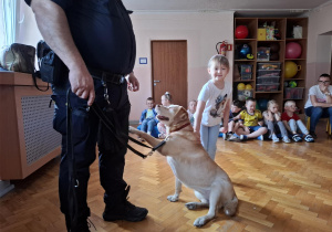 dziewczynka głaszcze psa w obecności strażnika miejskiego, na sali gimnastycznej w tle widać siedzące dzieci