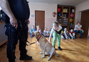 chłopiec głaszcze psa w obecności strażnika miejskiego, na sali gimnastycznej w tle widać siedzące dzieci