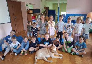 grupa dzieci na sali gimnastycznej pozuje do zdjęcia grupowego, przed nimi leży duży pies