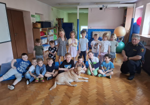 grupa dzieci na sali gimnastycznej pozuje do zdjęcia grupowego, przed nimi leży duży pies, z boku stoi przedstawiciel straży miejskiej