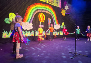 dzieci w kolorowych strojach występują na festiwalu na tle tęczy, prezentują piosenkę
