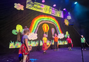 dzieci w kolorowych strojach występują na festiwalu na tle tęczy, prezentują piosenkę