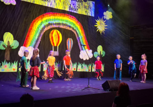 dzieci w kolorowych strojach występują na festiwalu na tle tęczy, prezentują piosenkę