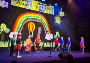 dzieci w kolorowych strojach występują na festiwalu na tle tęczy, prezentują piosenkę