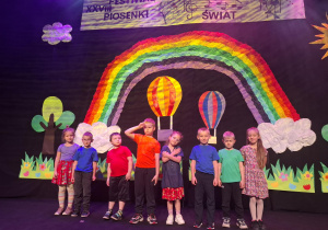 dzieci w kolorowych strojach występują na festiwalu na tle tęczy, prezentują piosenkę