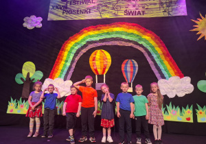 dzieci w kolorowych strojach występują na festiwalu na tle tęczy, prezentują piosenkę