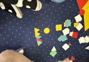 Dzieci siedzą na dywanie i układają kompozycje z figur geometrycznych.