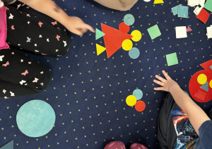 Dzieci siedzą na dywanie i układają kompozycje z figur geometrycznych.