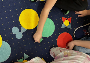 Dzieci siedzą na dywanie i układają kompozycje z figur geometrycznych.