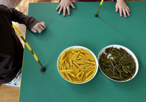 Dzieci siedzą przy stole i nawlekają makaron rurki na spaghetti.