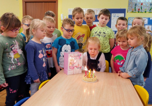 dziewczynka dmucha świeczki na torcie, dzieci stoją przy stoliku