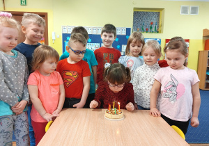 Grupa dzieci stoi przy stoliku, dziewczynka dmucha świeczkę na torcie.