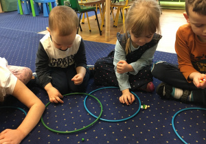 Dzieci siedzą na dywanie w ułożonych przed sobą kołach układają odpowiednią liczbę kolralików.