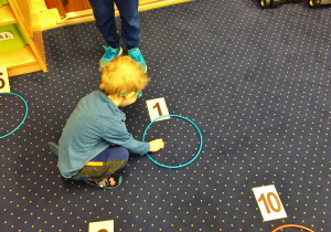 W ułożonych na dywanie obręczach chłopiec wkłada odpowiednią liczbę koralików do koła.