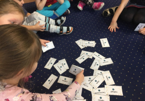 Dzieci siedzą na dywanie pośrodku leżą karteczki z różnymi słowami, które dzieci wybierają.