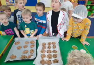 Dzieci stoja przy stole na którym leżą blach do pieczenia z gotowymi do tego procesu ciasteczkami owsianymi.