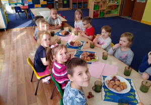 Przy dwóch stolikach siedzą dzieci. Na stołach znajdują się talerze z pączkami i winogronem