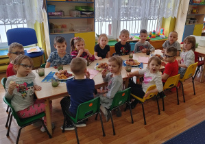 Przy dwóch stolikach siedzą dziewczynki i chłopcy. Na stołach kolorowe serwetki, na talerzach pączki i winogrono.