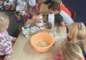 Dzieci siedza przy stolei obserwują jak pani odmierza na wadze produkty do ciasteczek.