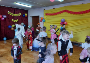 W tle dekoracja. Po prawej żółty materiał ozdobiony balonami, po lewej bordowy materiał z napisem "Karnawał 2021". Na pierwszym planie dzieci tańczą do muzyki.