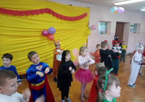 Po lewej stronie widać ścianę ozdobioną żółtą tkaniną i balonami. Grupa dzieci w przebraniach karnawałowych tańczy.