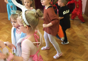 Dzieci w przebraniach, w kole tańczą do muzyki powtarzając ruchy prezentowane przez nauczyciela.