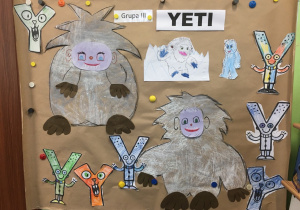 Praca plastyczna dzieci. Na szarym kartonie dwie postaci "Yeti" wykonane pastelami z wykorzystaniem systemu EPR oraz wycięte, pokolorowane litery "y".