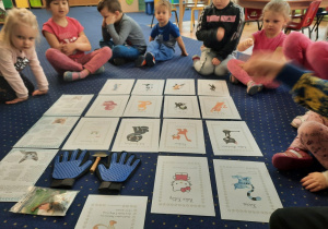 Dzieci siedzą w kole i nazywają postaci kota z bajek. Na podłodze ilustracje kotów z bajek oraz przyborów do higieny i zabawy z kotem.