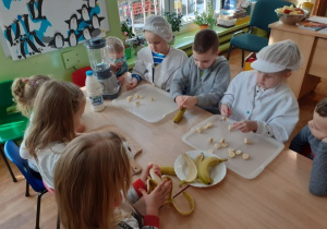 Dzieci siedzą przy stołach. Dwie dziewczynki kroją banany, a dziewczynka z chłopcem obiera banany ze skórki.